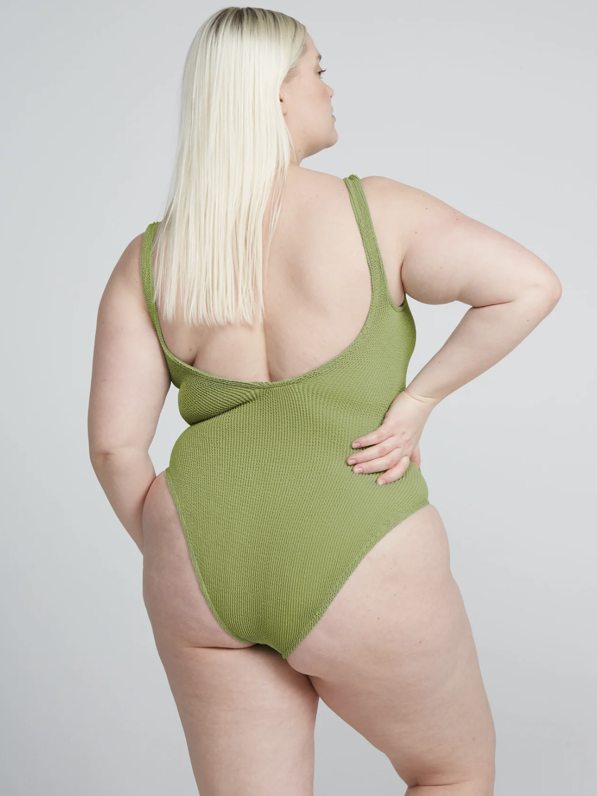 Person with long blonde hair wearing a green ribbed one-piece swimsuit stands facing away, with hand on hip against a neutral background.