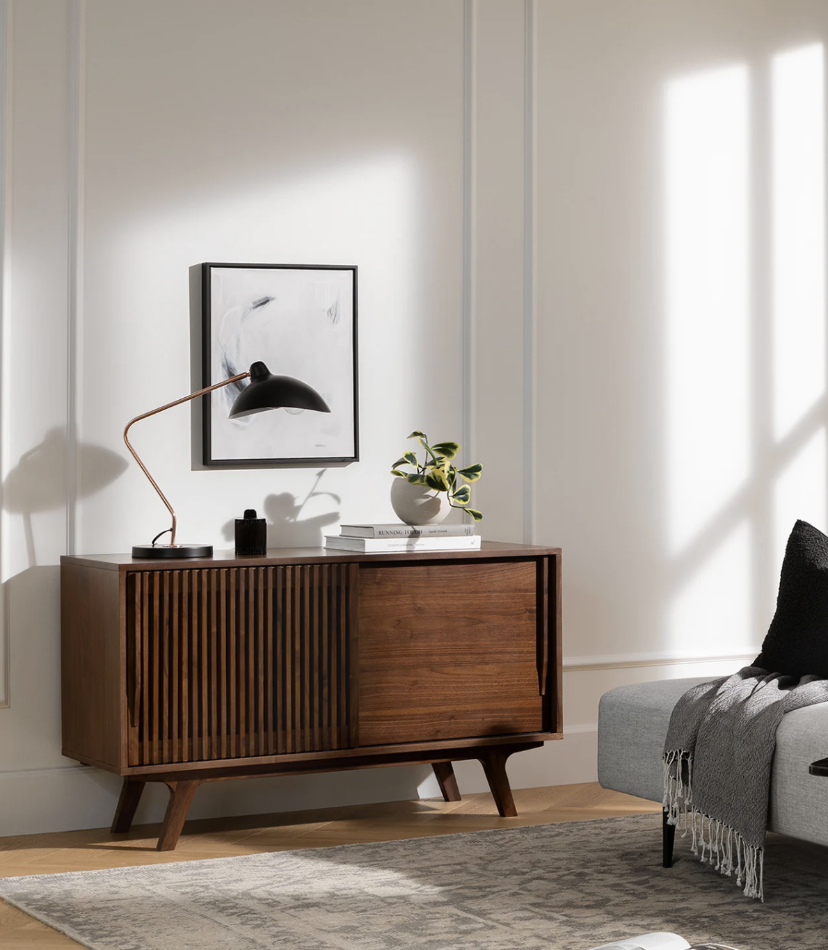A modern wooden sideboard with a lamp, plant, and books on top, stands against a white wall with framed art, next to a gray sofa and a patterned rug.