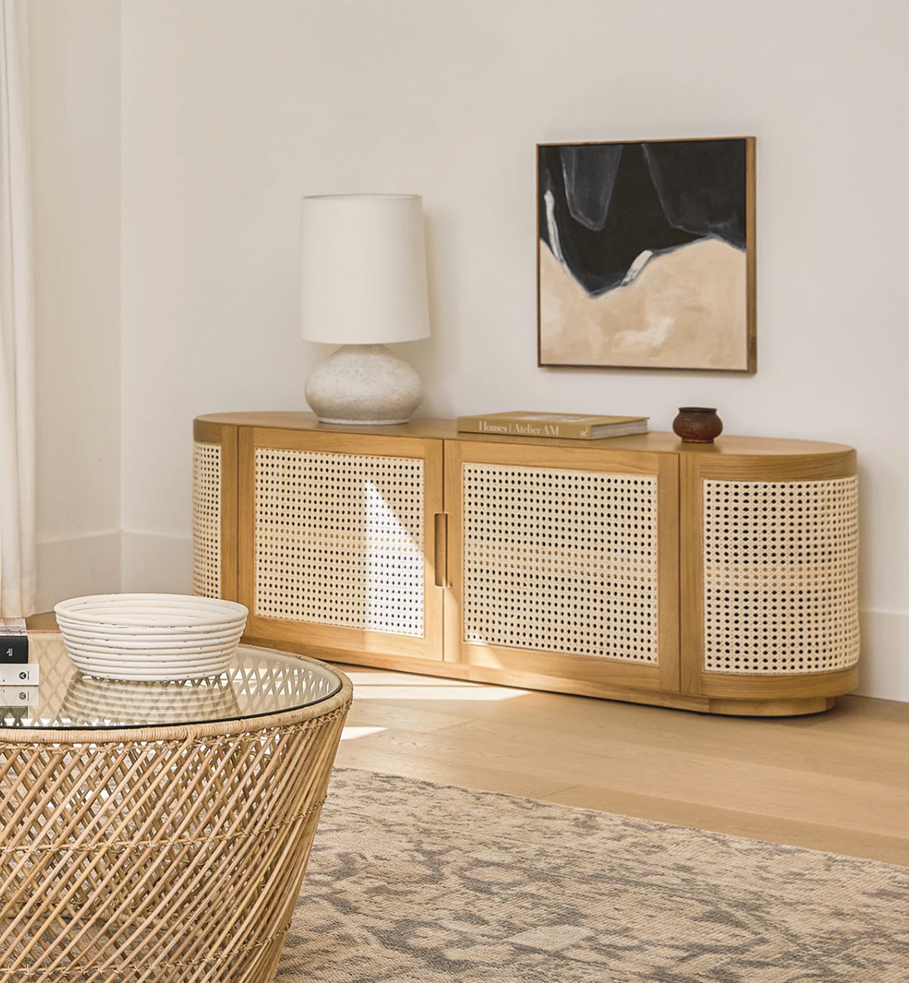 A light wood credenza with woven rattan doors, a white lamp, books, and a small bowl on top, sits against a white wall with abstract art above.