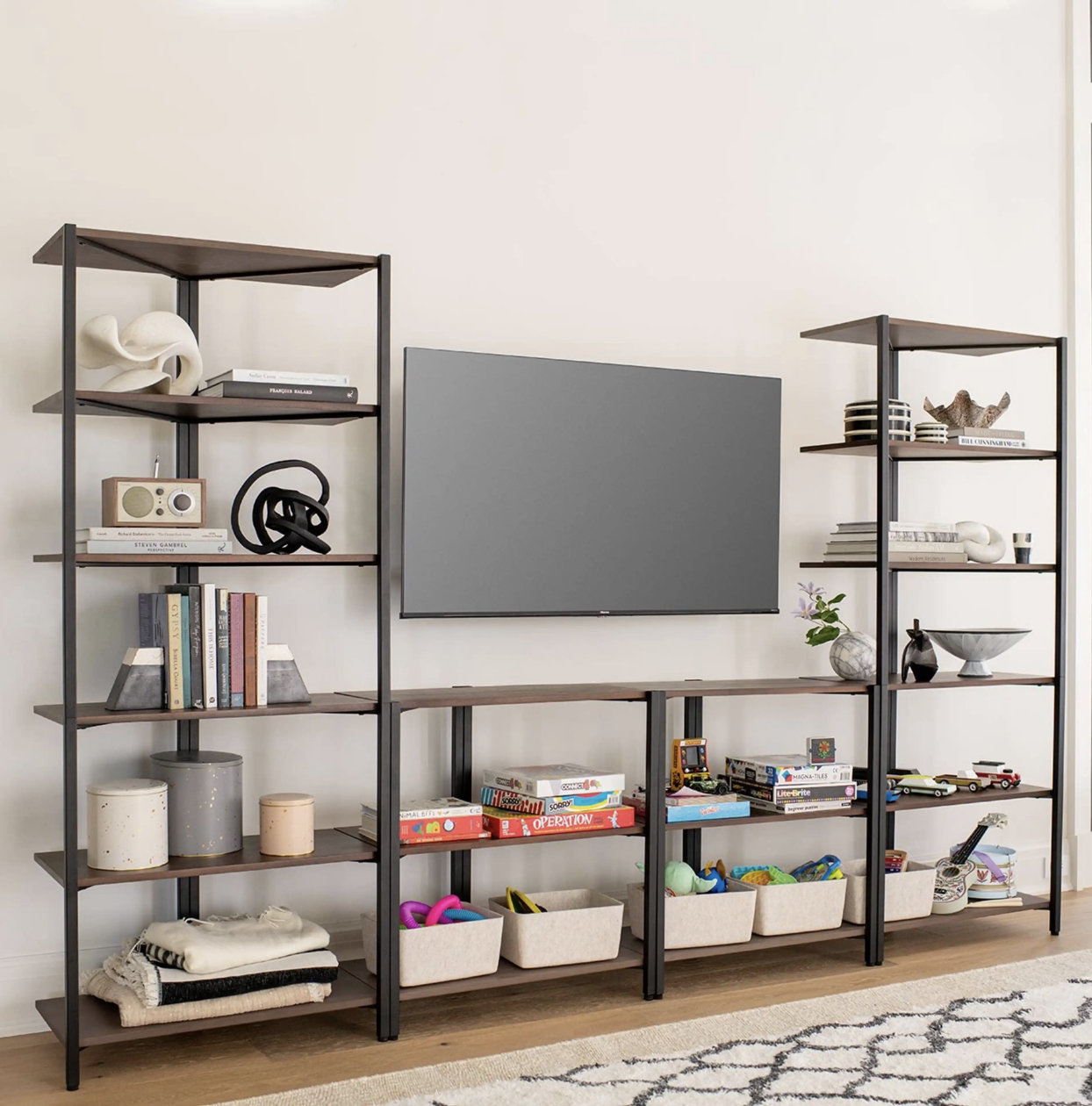 A TV mounted on a wall is flanked by open shelving units holding books, decor, plants, games, and storage bins with toys in a living room setting.