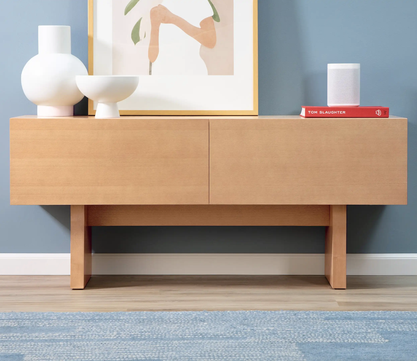 Light wood sideboard against a blue wall with abstract art above; two white decorative objects and two books on top; blue rug on the floor.