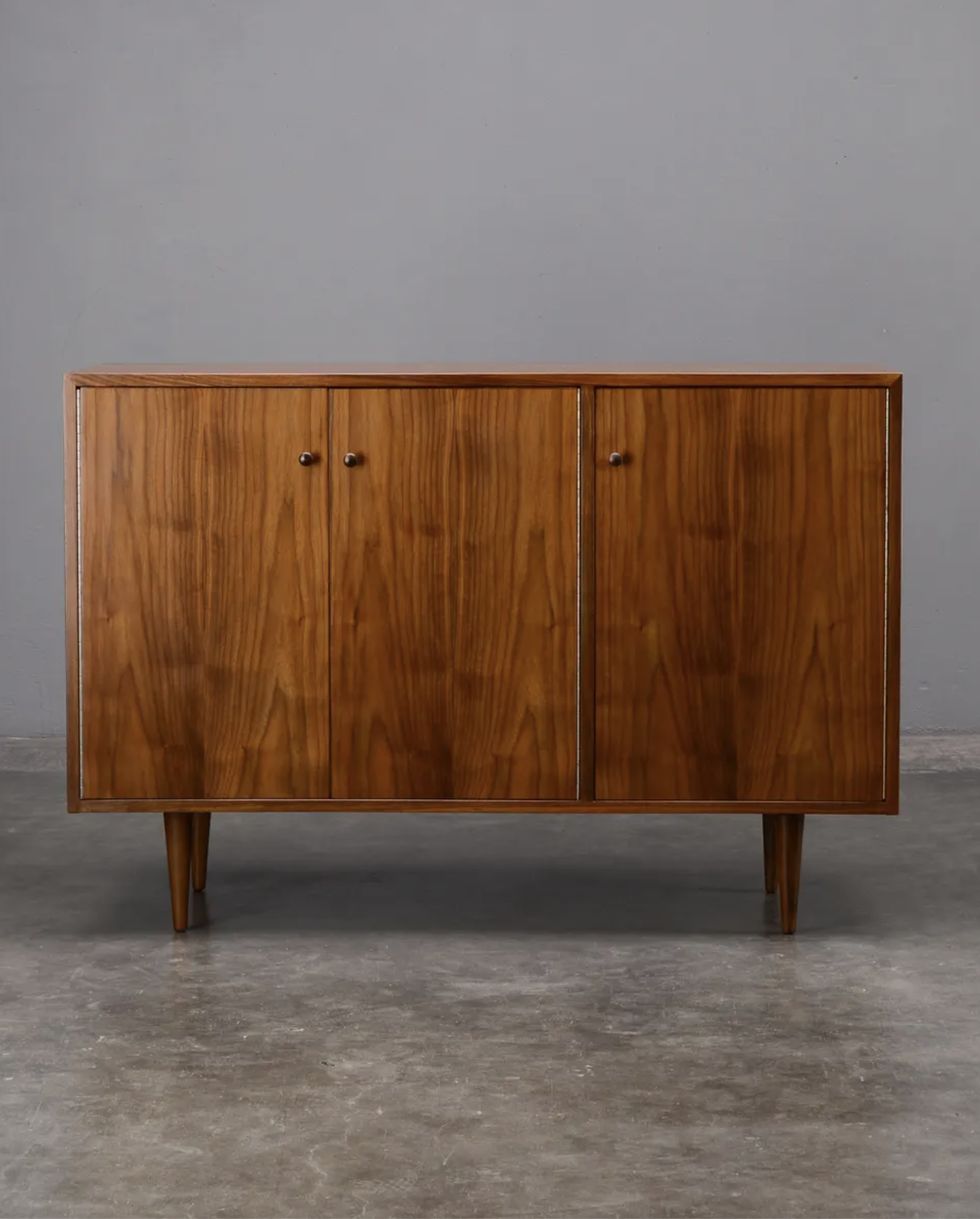 A mid-century modern wooden sideboard with three cabinet doors and tapered legs, set against a plain gray wall and concrete floor.