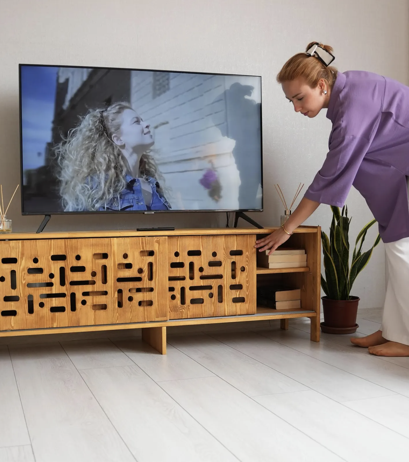 A woman in a purple top organizes items in a wooden TV console while a flat-screen television displays a woman outdoors. A potted plant is on the floor beside the console.
