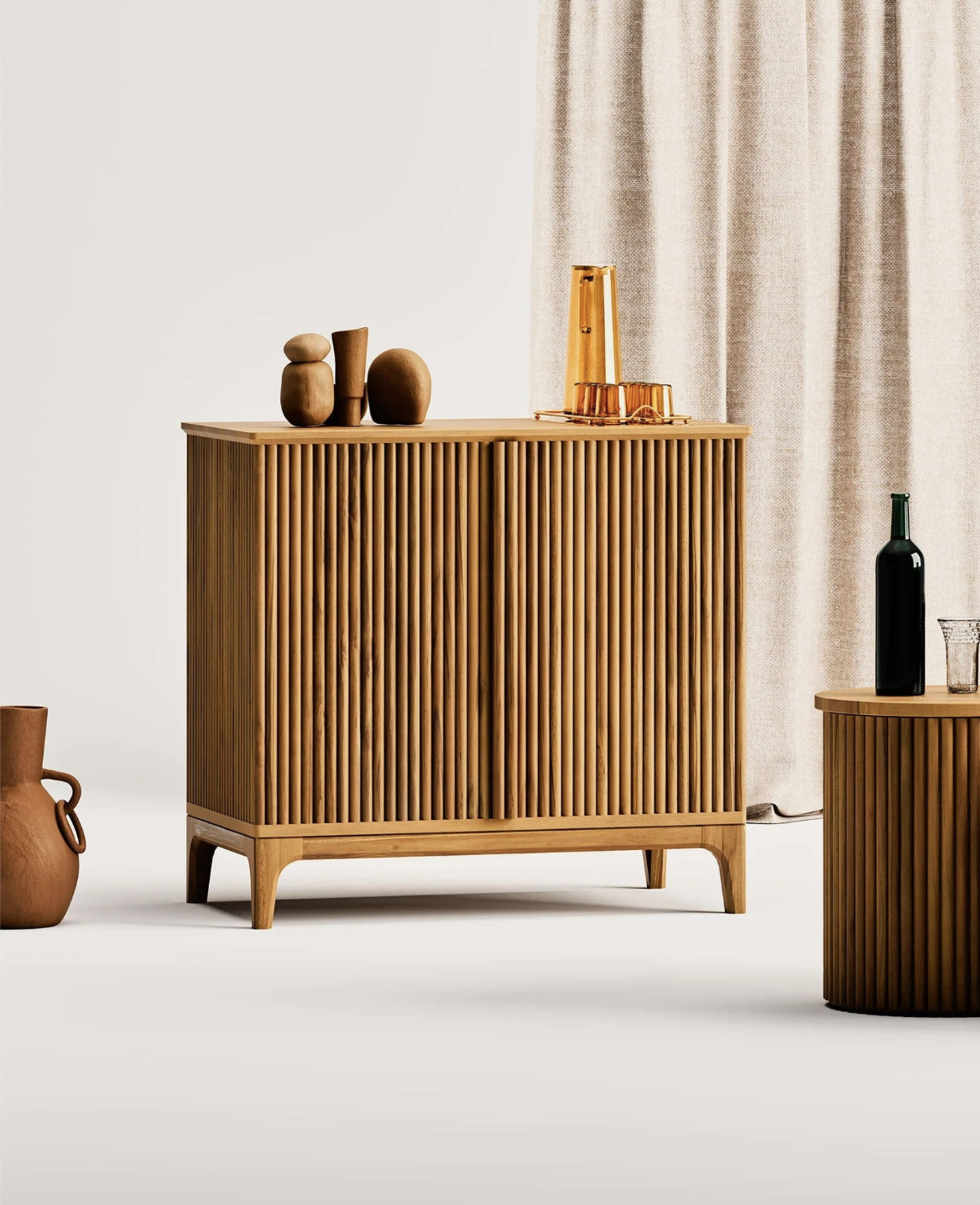 A wooden sideboard with vertical grooves stands against a light-colored curtain, topped with decorative objects, a carafe, and glasses. A matching table and vase are nearby.