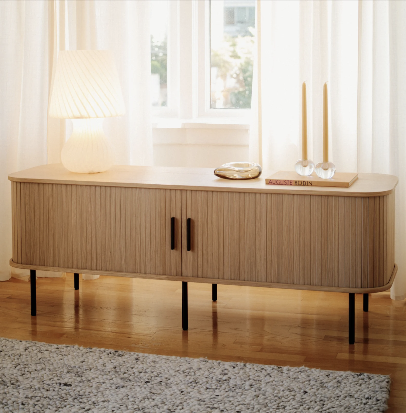 A wooden sideboard with vertical grooves, black legs, and two handles stands against a window. On top are a lamp, a book, a tray, and two candleholders.