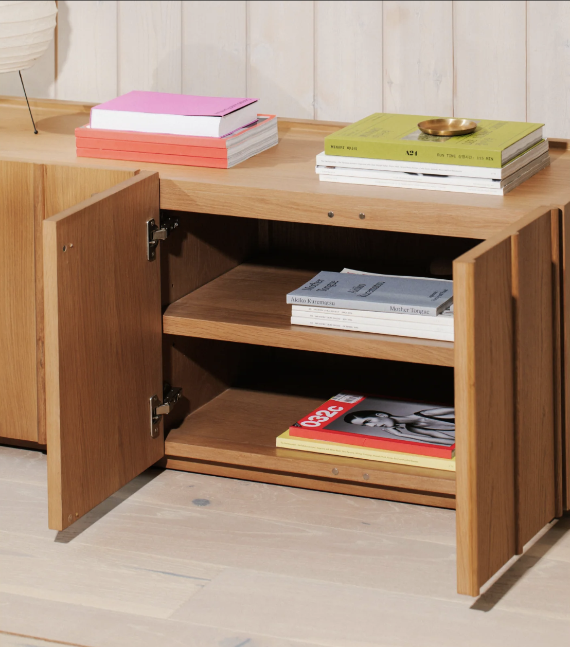 A wooden cabinet with two doors open, showing shelves inside holding magazines; books are stacked on top of the cabinet.