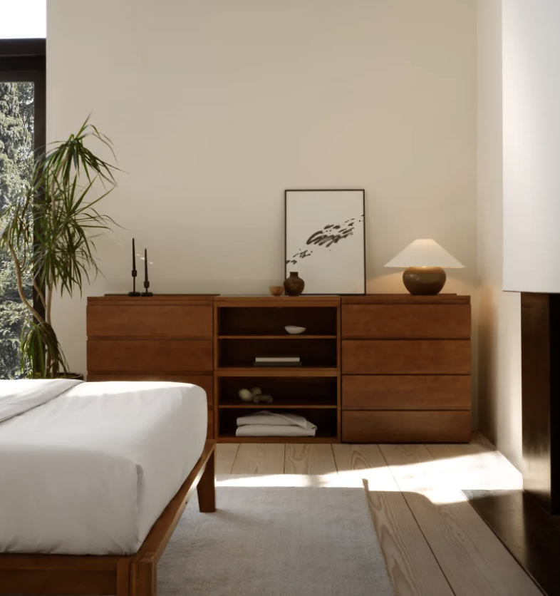 A minimalist bedroom features a wooden dresser with decor, including a lamp, framed art, and candles; a bed with white linens and a potted plant are also visible.