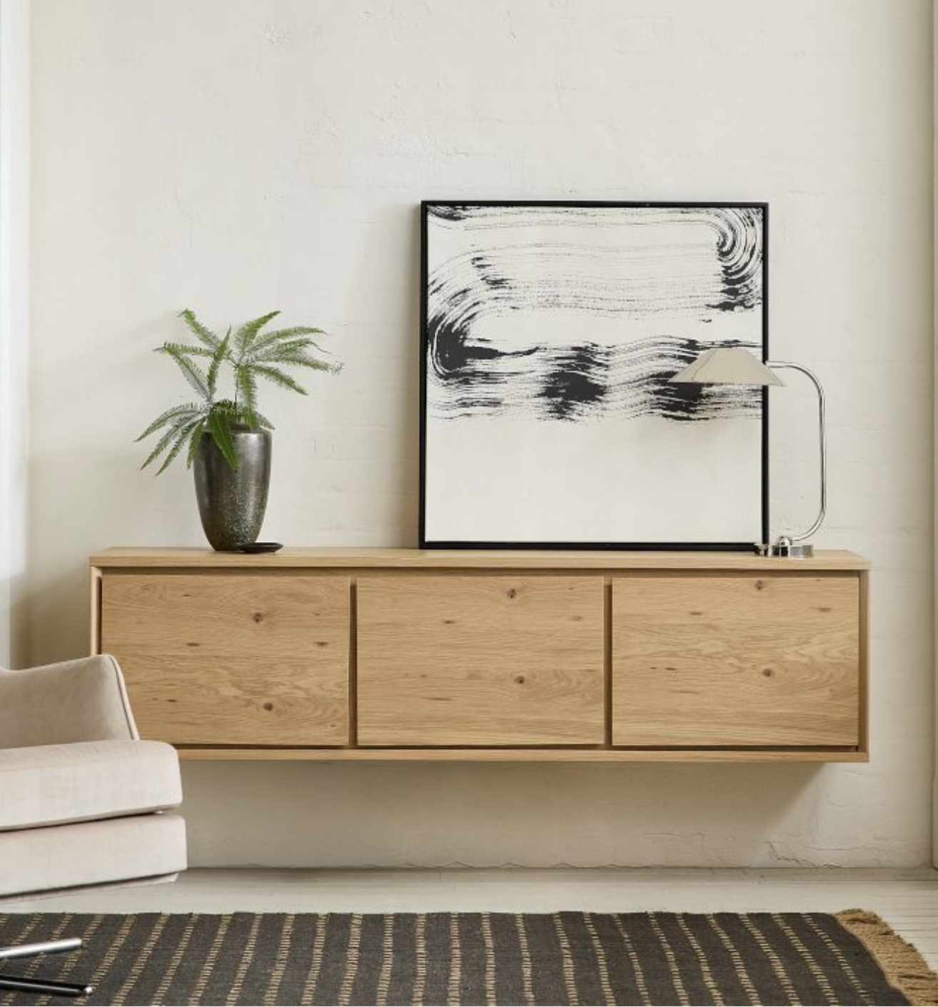 A minimalist wooden sideboard with three doors is mounted on a white wall, topped with a potted plant, abstract black-and-white artwork, and a modern desk lamp.