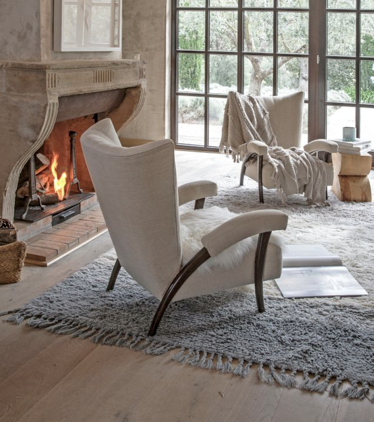 Cozy living room with a fireplace, two upholstered chairs, and a gray rug. A throw blanket is draped over one chair. Large windows reveal trees outside. An open book lies on the rug.
