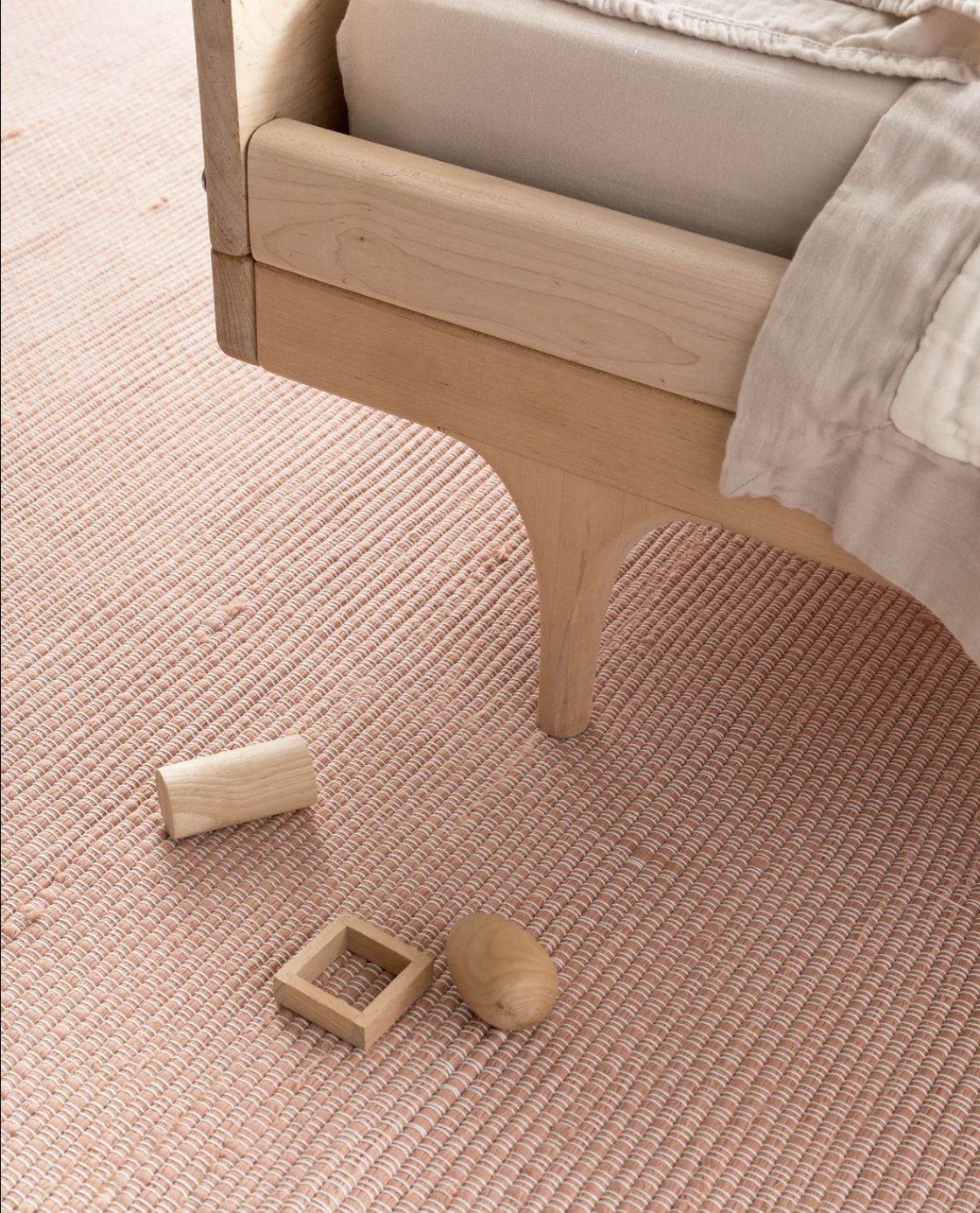 Wooden bed frame on a textured pink rug with three wooden toys: a cylinder, a cube with a square hole, and an egg-shaped piece nearby.