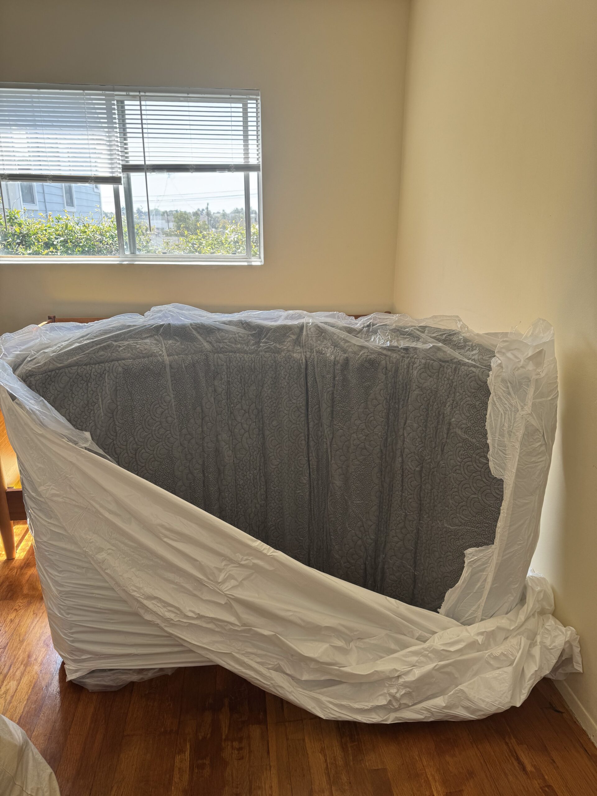 Unassembled furniture part wrapped in plastic sits in a room with wooden floors and large window blinds.