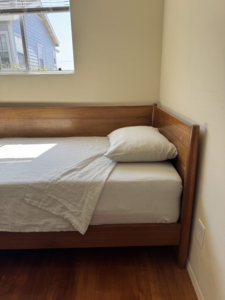 A neatly made wooden bed with white sheets and a pillow is positioned against a beige wall near a window with sunlight streaming in.