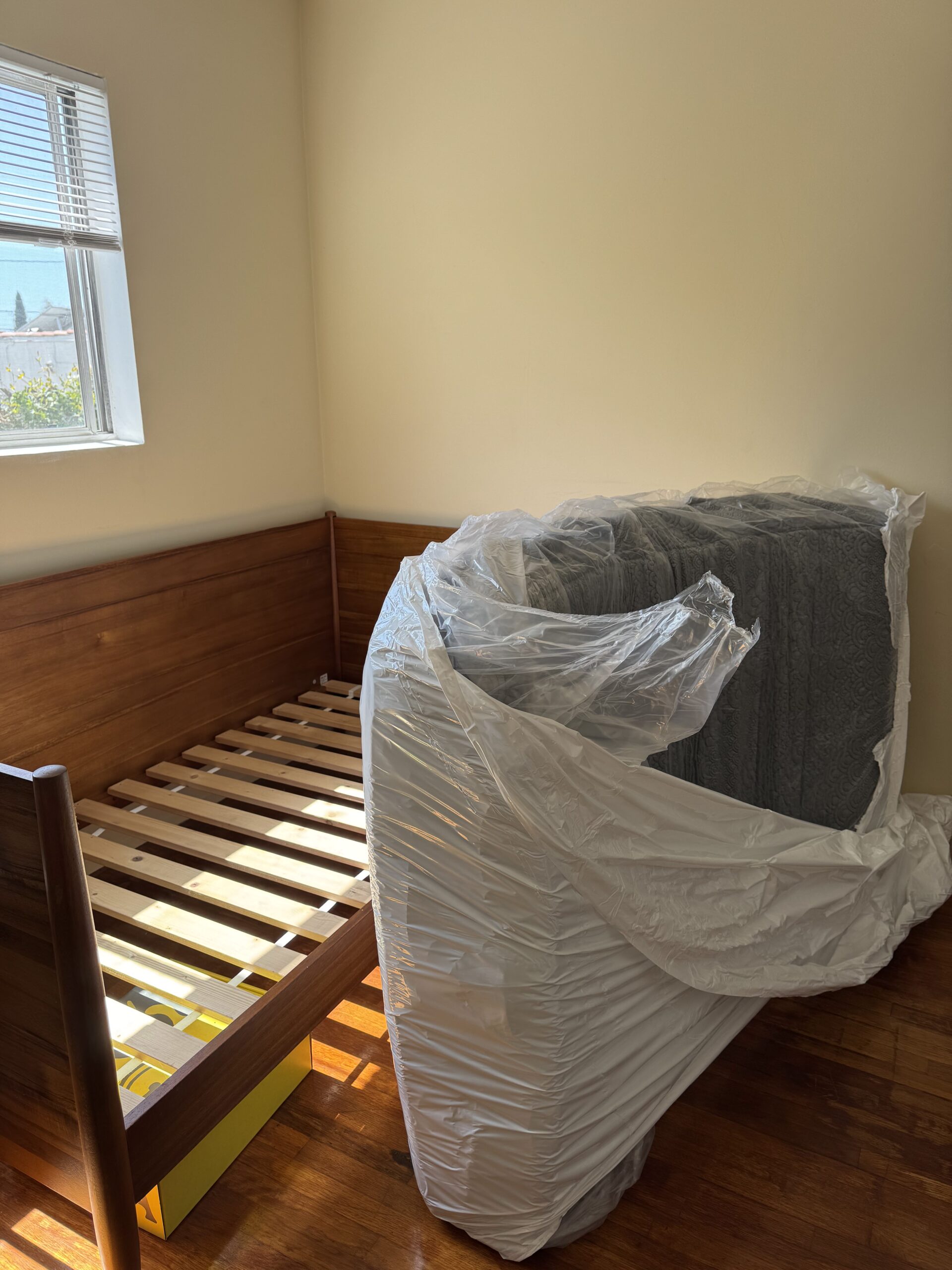 A wooden bed frame is assembled next to a window. A wrapped mattress stands upright beside it in a bright, empty room.