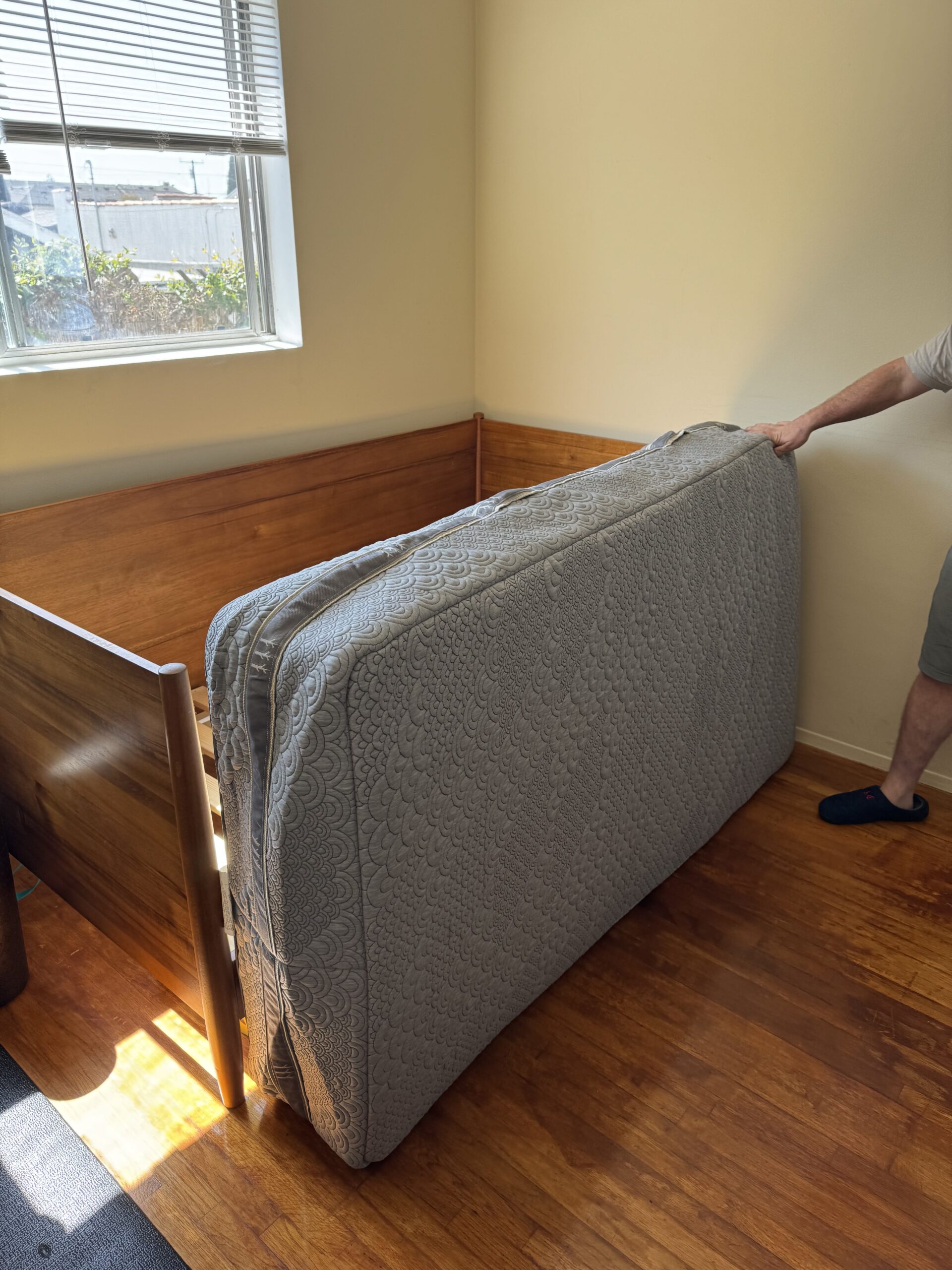 Person holding an upright mattress beside a wooden bed frame in a sunlit room.
