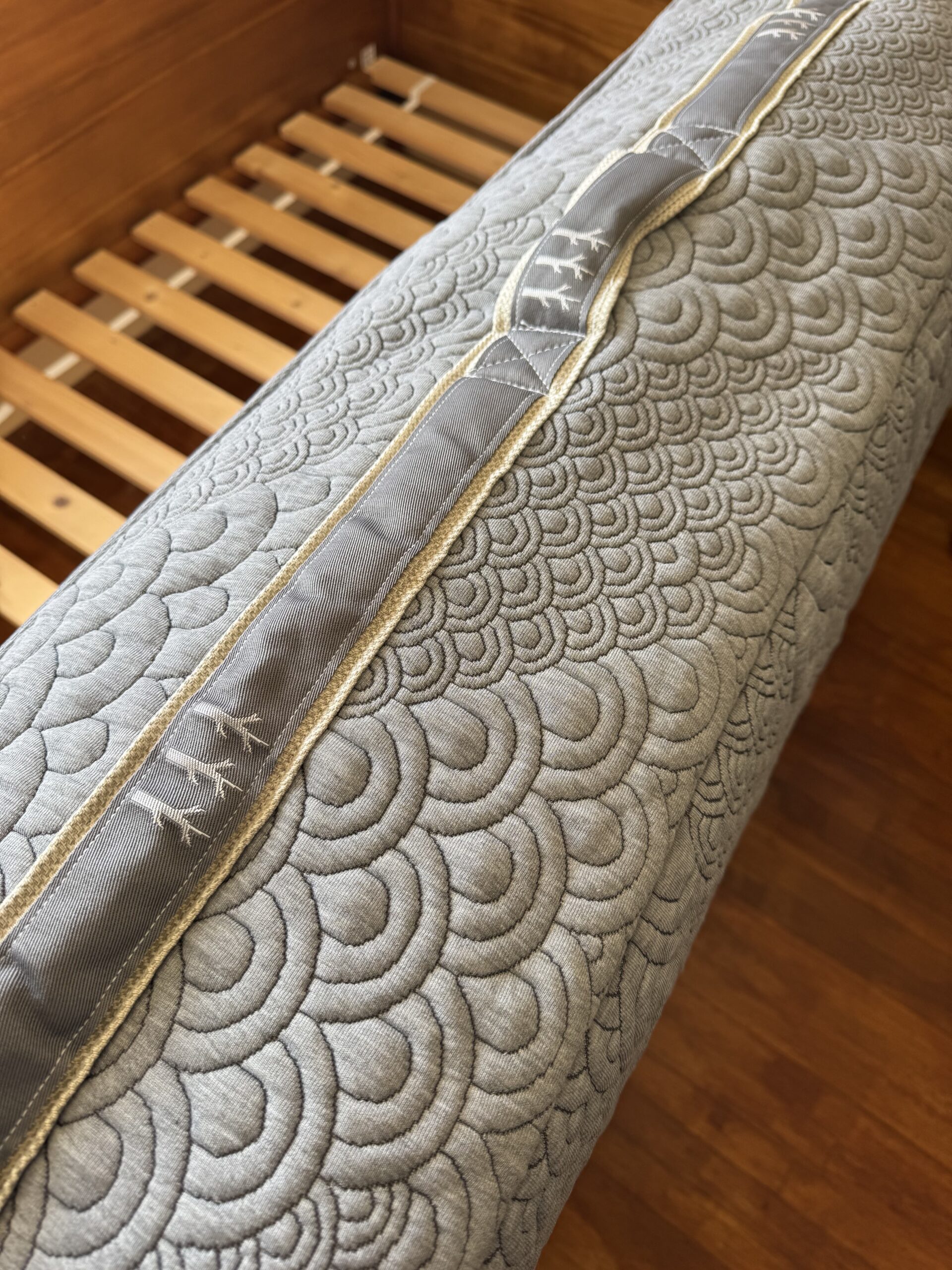 Close-up of a rolled-up mattress on a wooden bed frame, showing a gray quilted pattern with spiral designs and a stitched gray handle loop.