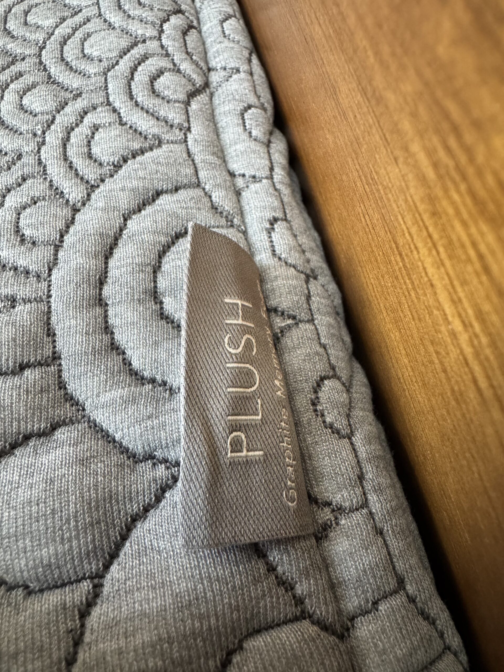 A close-up image of a gray, quilted fabric with swirling patterns, featuring a fabric tag labeled "PLUSH." The fabric is adjacent to a wooden surface.