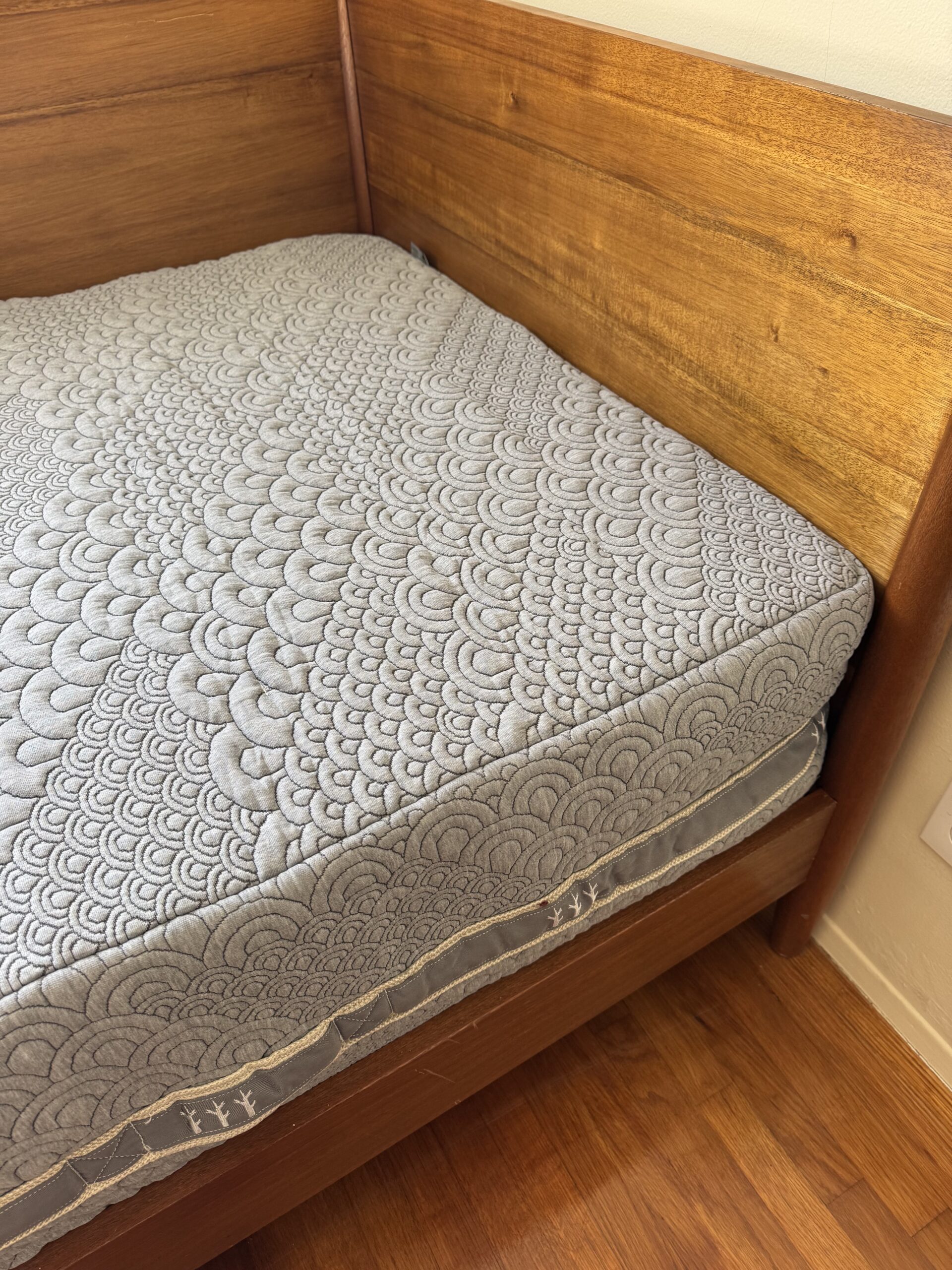 A wooden bed frame with a mattress covered in a textured, patterned gray fabric. The corner of the bed is visible, showing a hardwood floor surface.