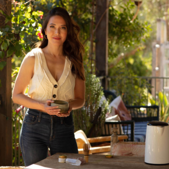 A woman in a sleeveless top holds a bowl while standing beside a table outdoors, surrounded by greenery and sunlight.