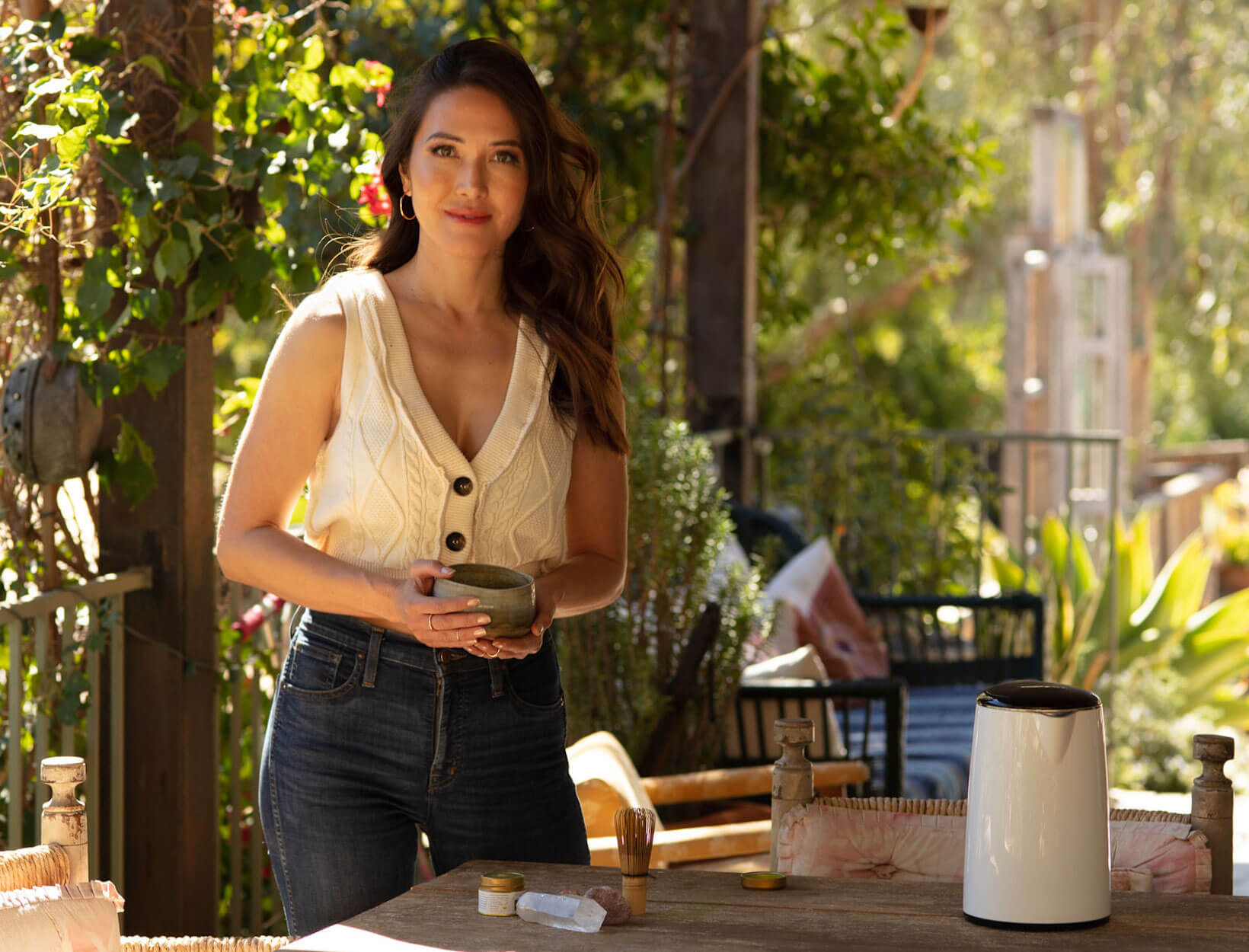 A woman in a sleeveless top holds a bowl while standing beside a table outdoors, surrounded by greenery and sunlight.