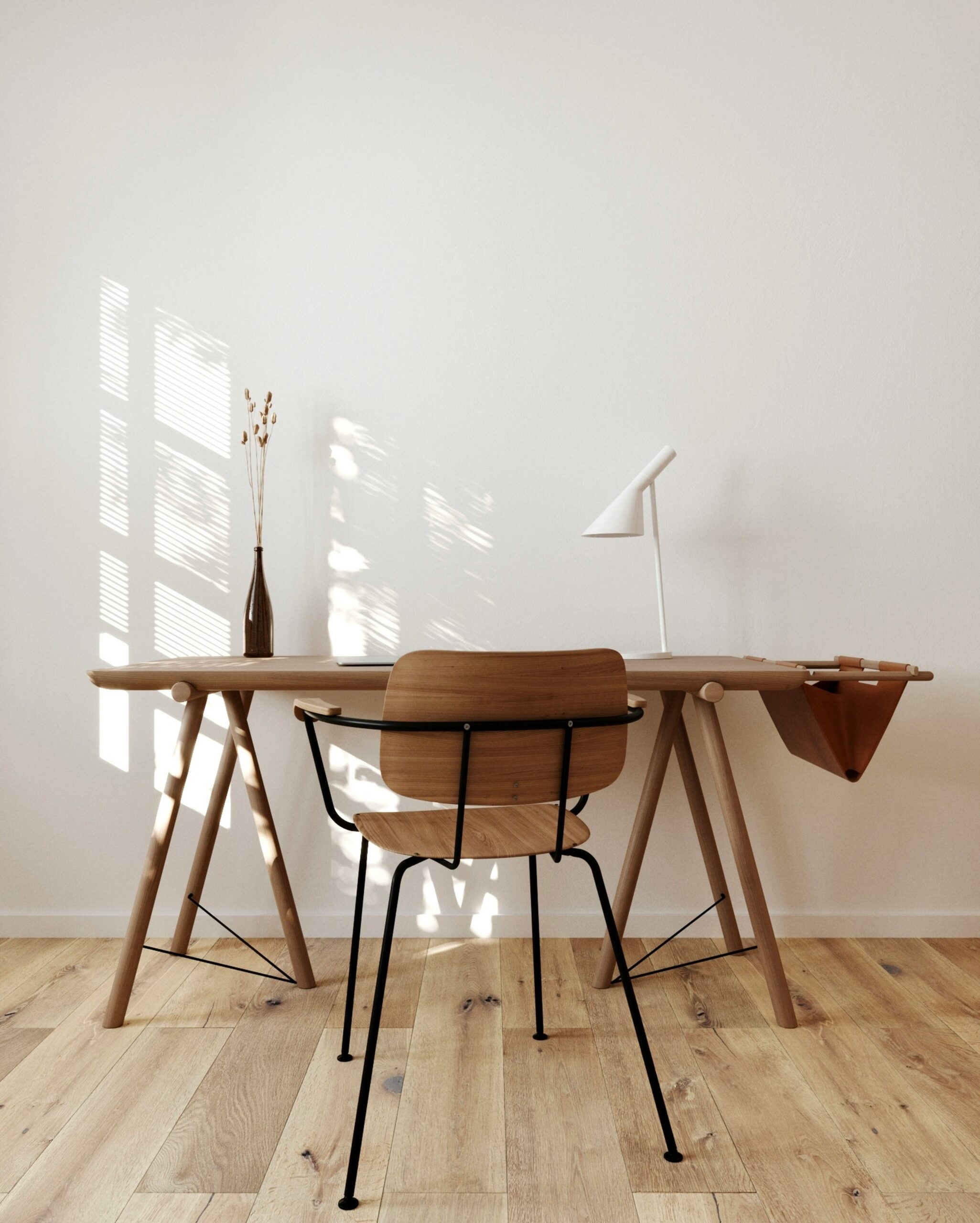 Minimalist workspace with a wooden desk, black chair, desk lamp, and a vase with dried flowers. Sunlight creates patterns on the wall and floor.