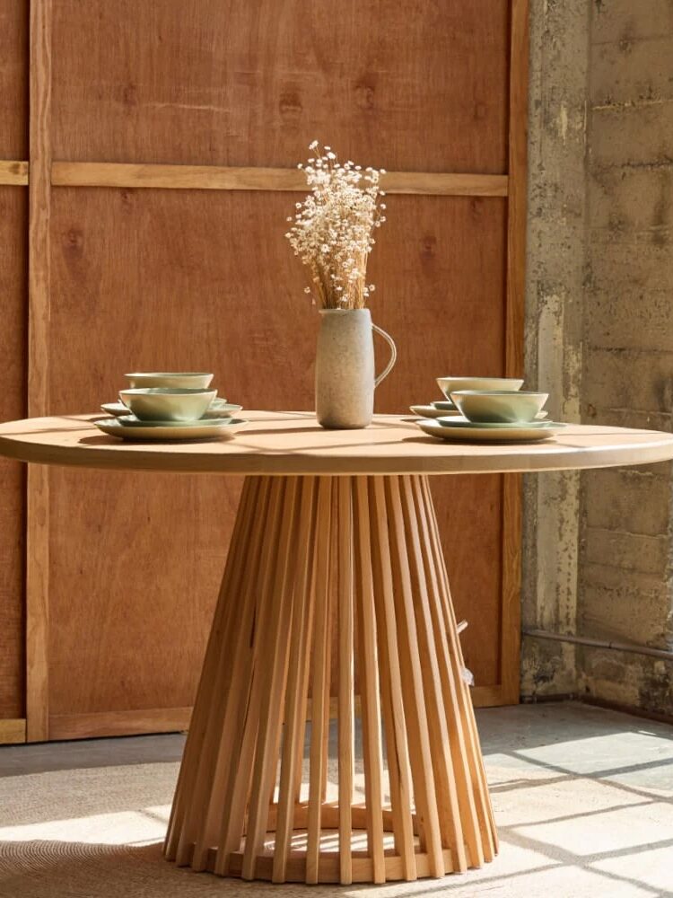 A round wooden table with a slatted base, set with four place settings and a ceramic jug of dried flowers, stands near a large window in a sunlit room.