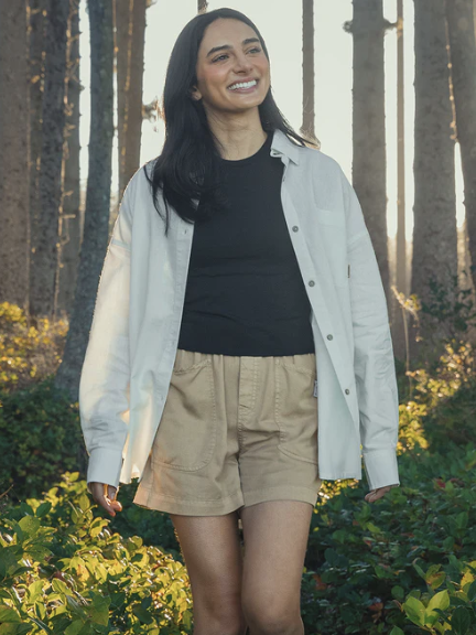 A woman wearing a black shirt, beige shorts, and an open light-colored button-up shirt walks outdoors in a sunlit forest.