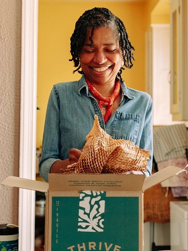 A woman stands in a kitchen smiling while unpacking items from a Thrive Market box. Various groceries are visible on the counter in front of her.