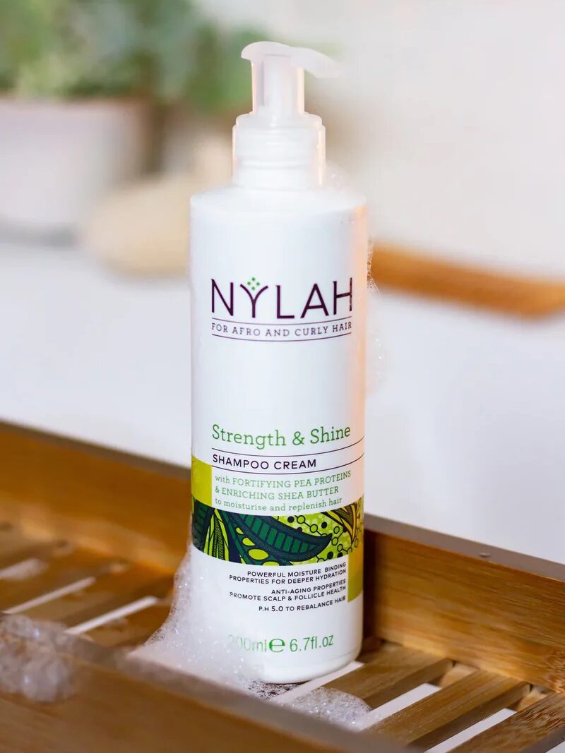 A bottle of Nylah Strength & Shine shampoo cream for afro and curly hair on a wooden surface, surrounded by soap bubbles.