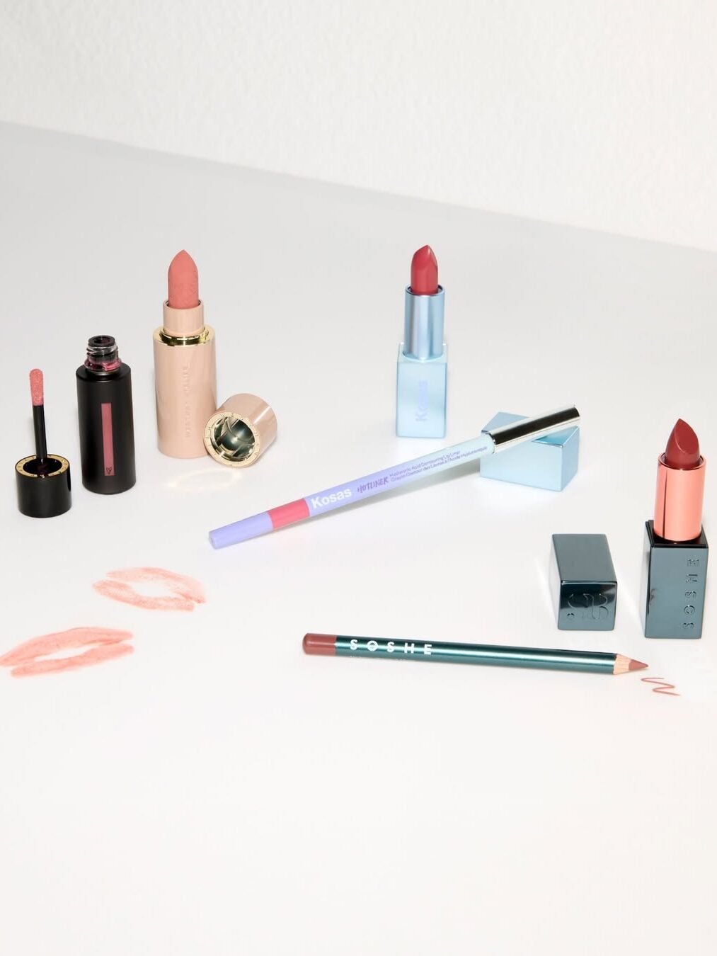 Various lipsticks, a lip liner, and lip gloss are arranged on a white surface. Lip imprints in pink and red are visible nearby.