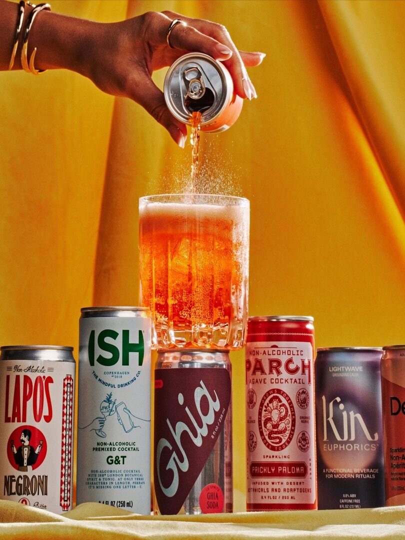 A hand pours a drink from a can into a glass against a yellow backdrop. Five different cans are displayed in the foreground.