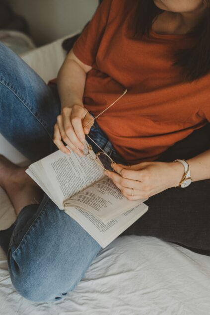 A person in a rust-colored shirt and jeans sits cross-legged on a bed, holding glasses and reading an open book.
