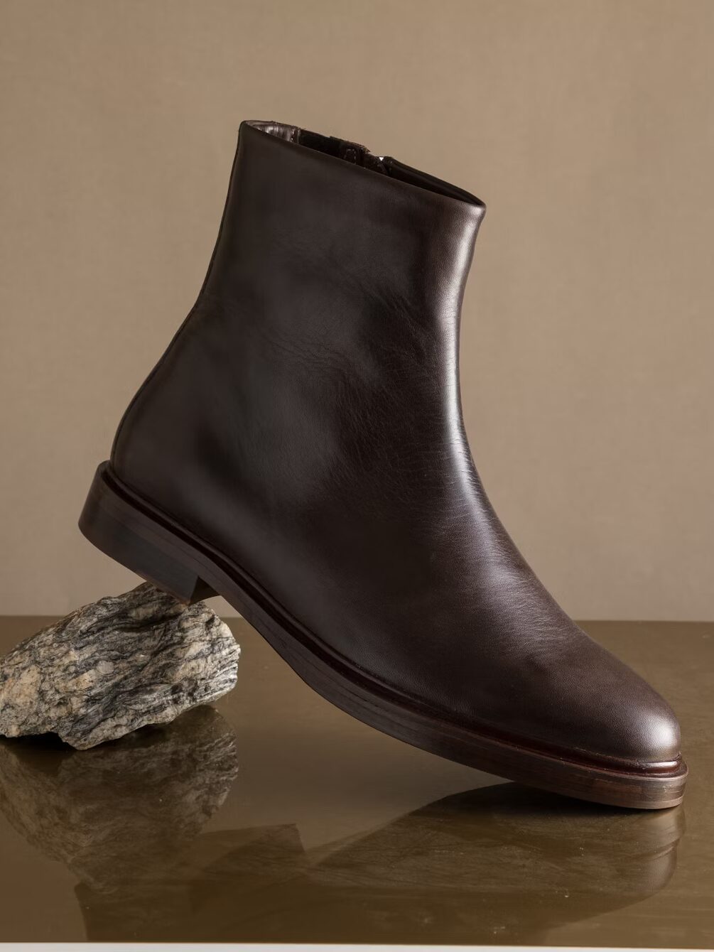 A dark brown leather ankle boot is balanced on a textured rock against a plain beige background.