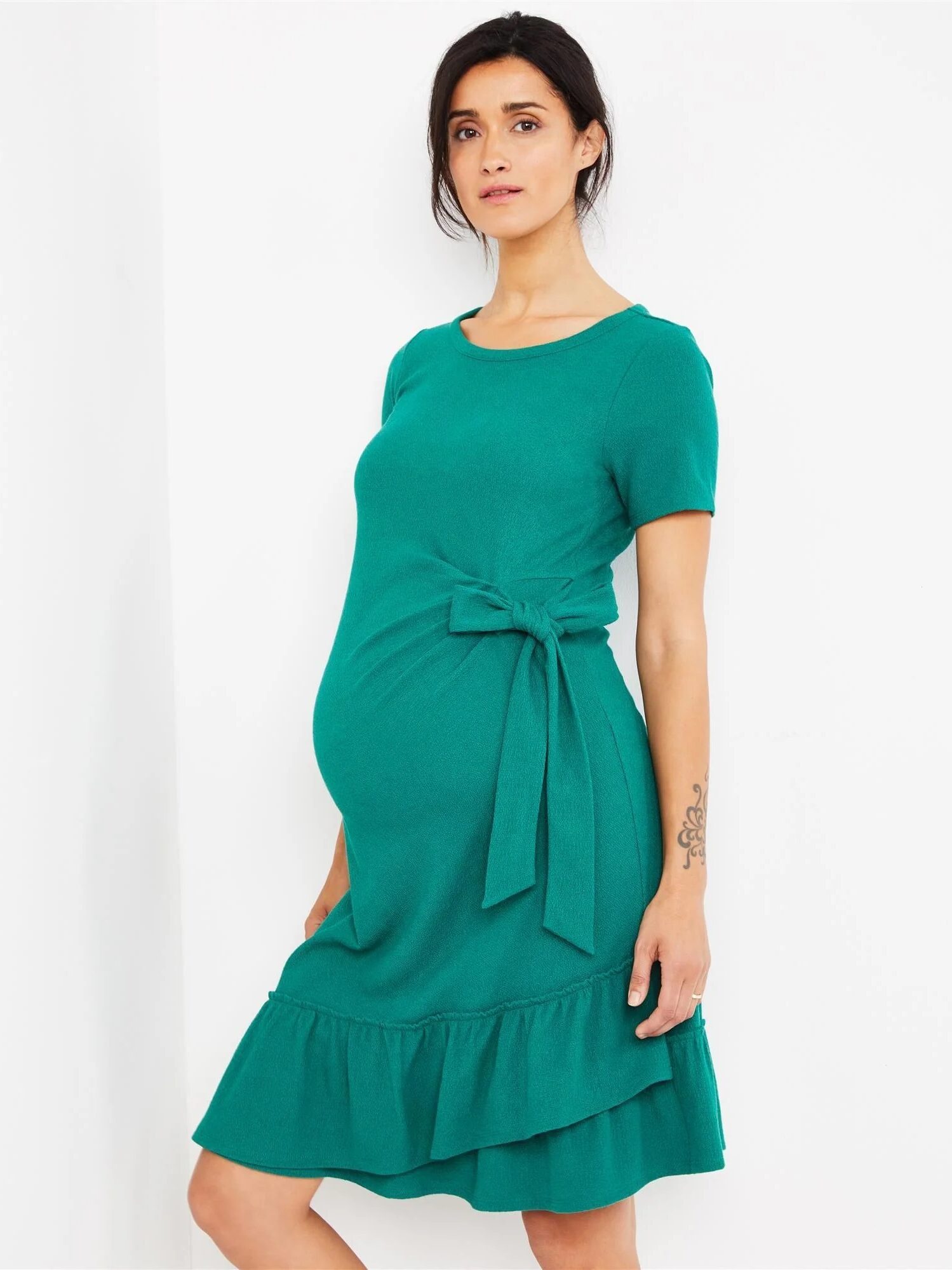 A woman wearing a green, short-sleeved maternity dress with a waist tie and ruffled hem, standing against a plain white background.