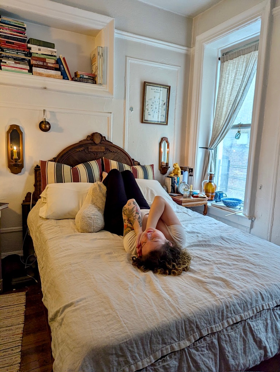 A person with curly hair lies on a bed in a cozy, sunlit bedroom with books on a shelf, plush pillows, and a large window with partially drawn curtains.