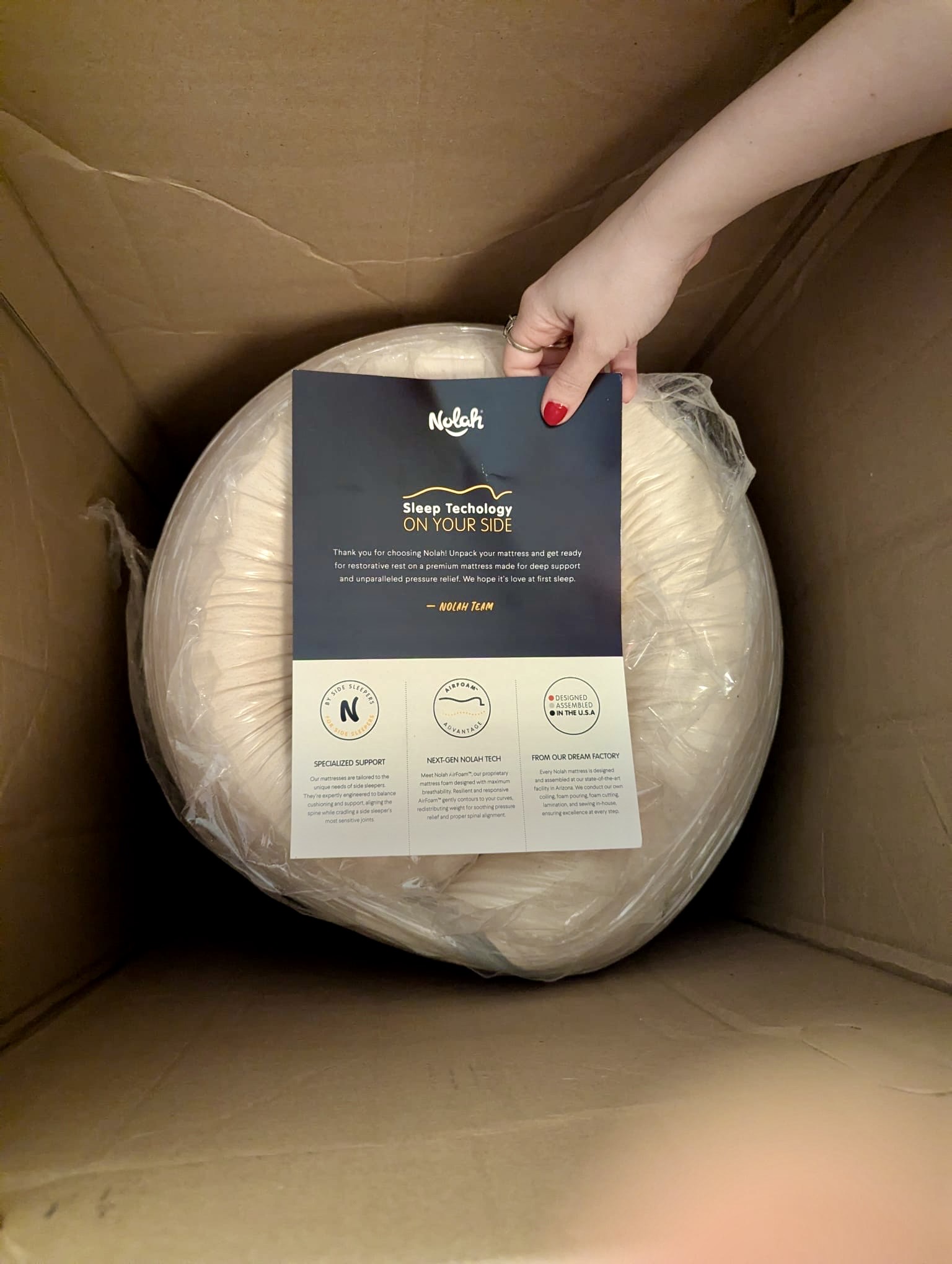 A hand holds an information card over a round, wrapped item inside a cardboard box. The card features the Nolah brand and details about sleep technology.