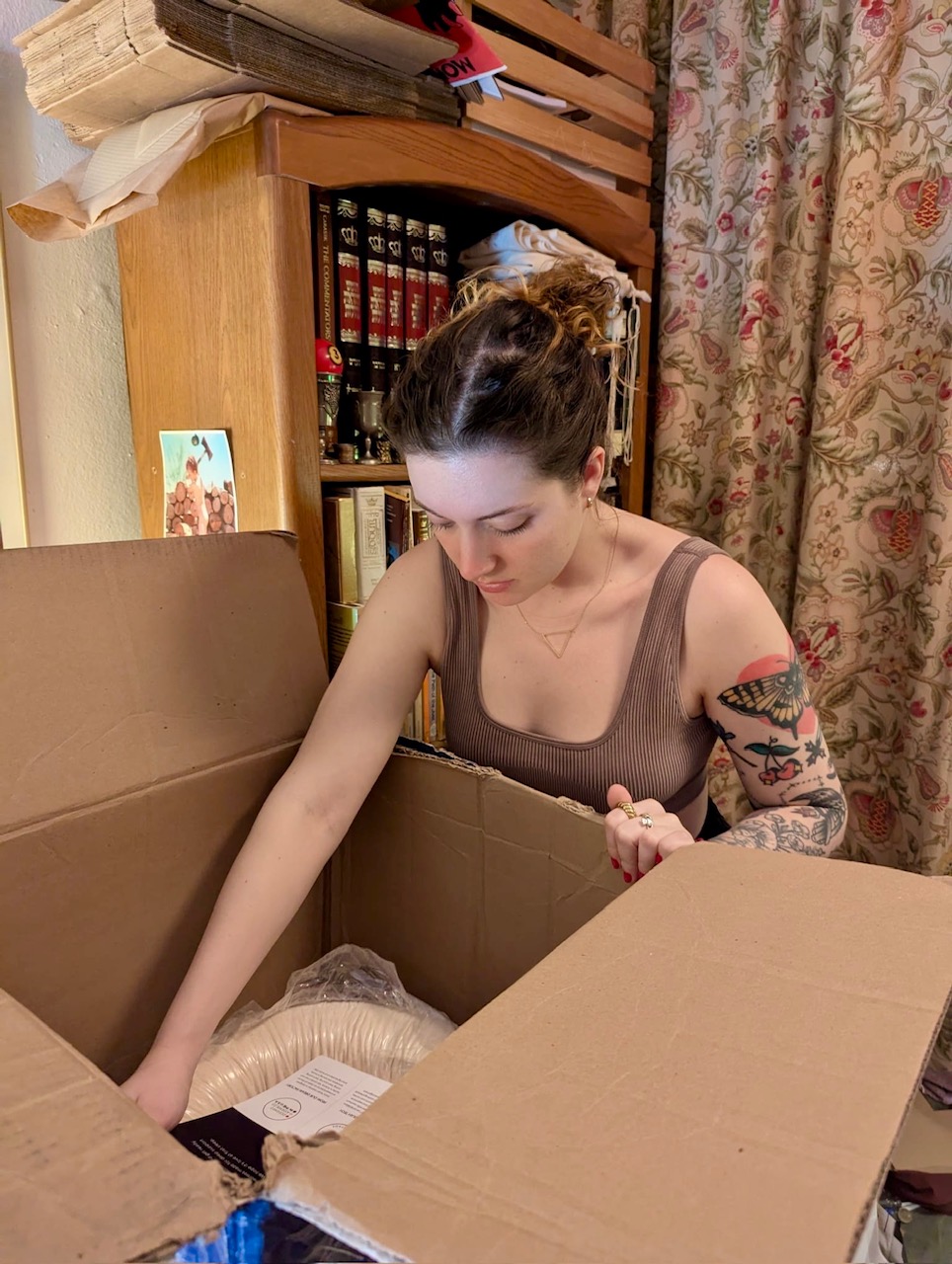 A woman with tattoos opens a large cardboard box in a room with bookshelves, floral curtains, and stacked items in the background.