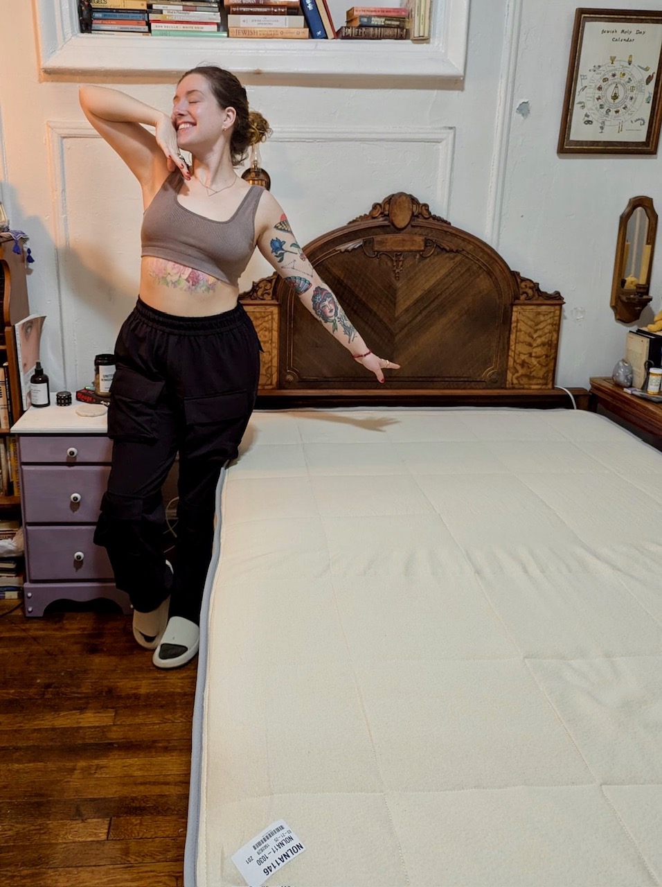 A person with tattoos stands and poses happily next to a neatly made bed with a visible mattress tag in a decorated bedroom.