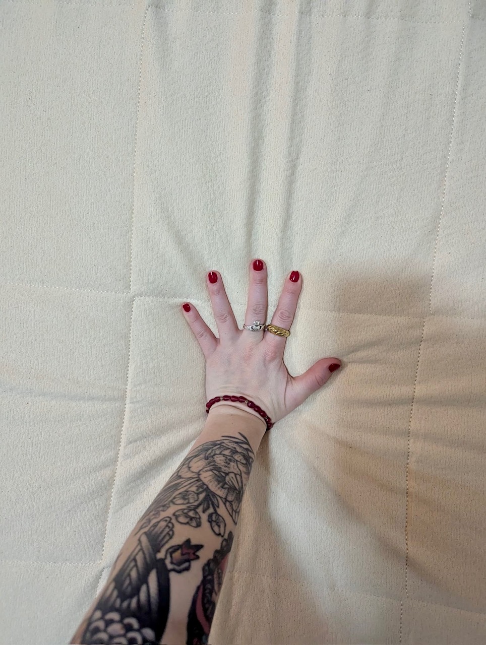 A tattooed arm with red nail polish and rings presses a hand into a beige quilted surface, creating an indentation.