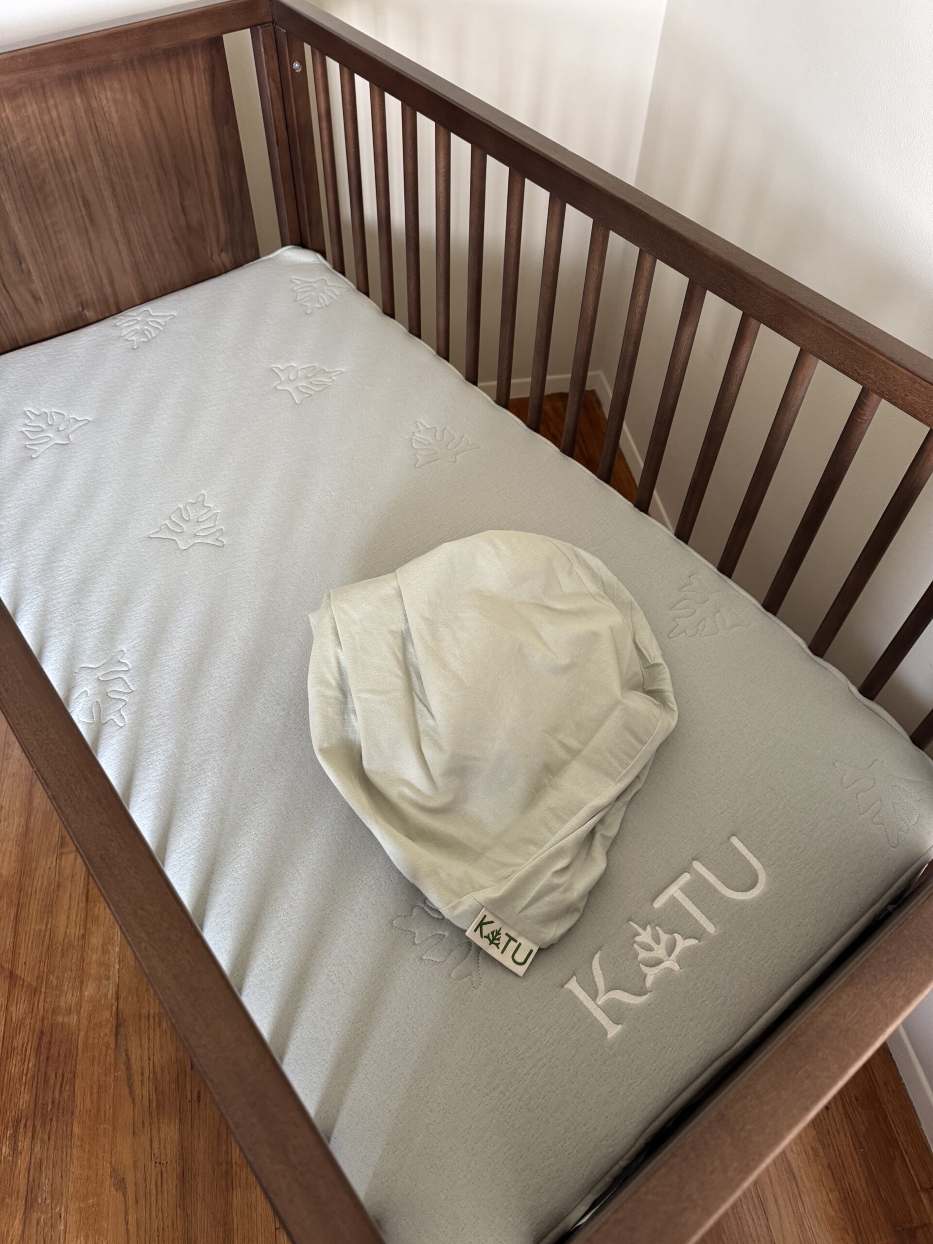 A wooden crib with a light gray mattress and a matching fabric item labeled "KATU" placed on top.