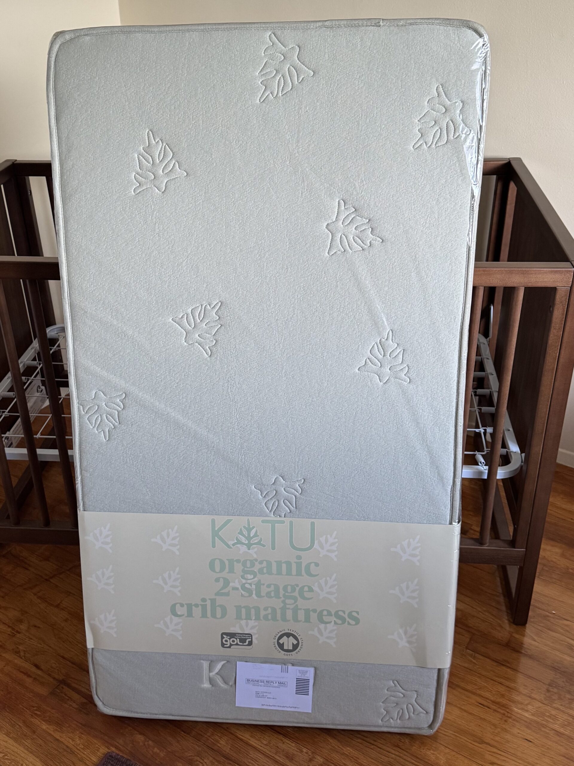 A new crib mattress stands upright in front of a wooden crib in a room with light-colored walls and a wood floor.