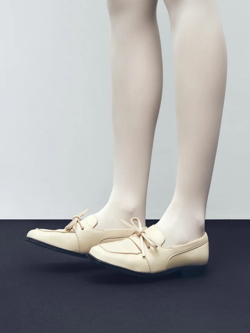 A person wearing white tights and beige loafers with a bow, standing on a black surface against a light gray background.