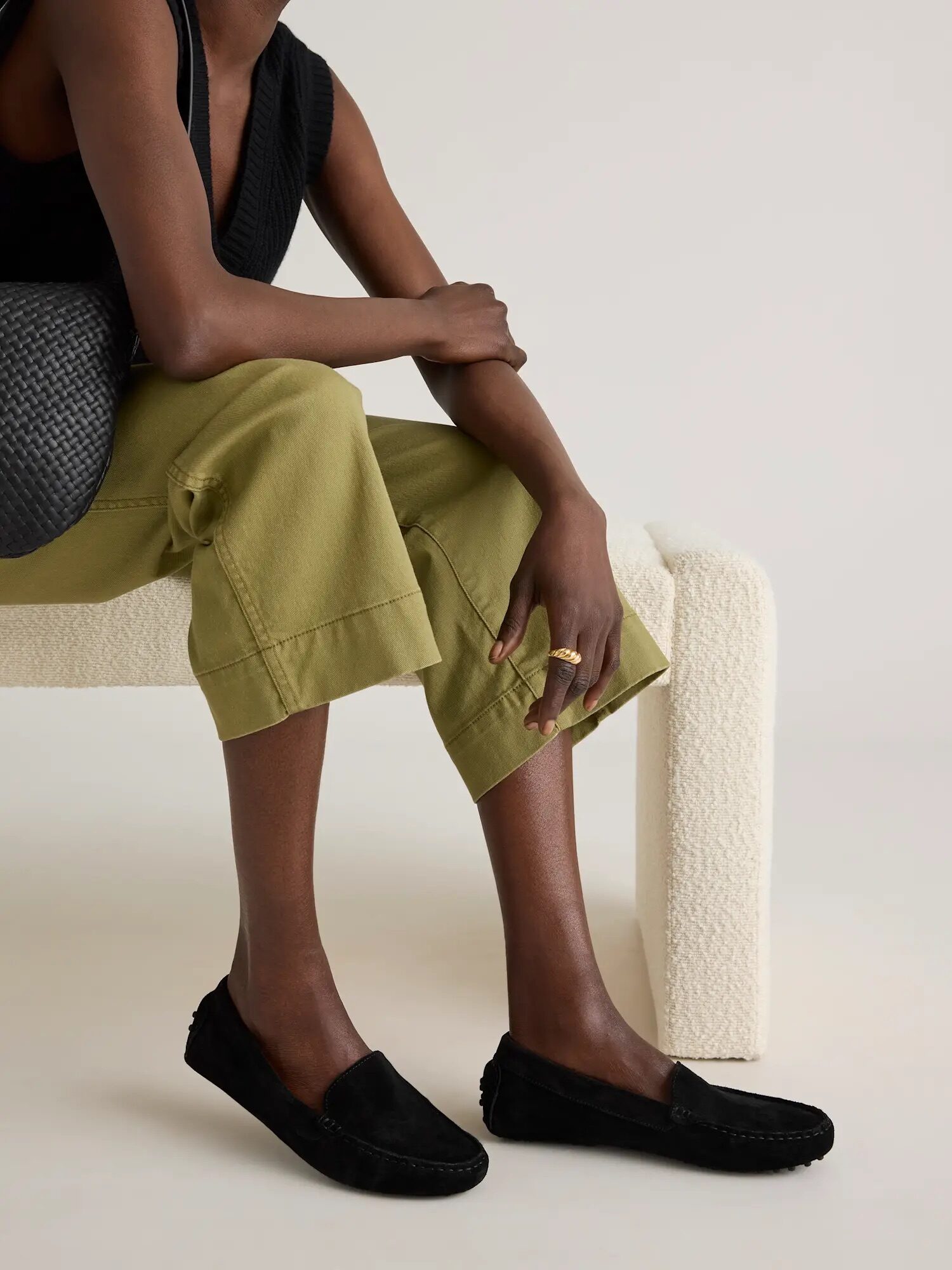 Person seated on a white bench wearing a black sleeveless top, light green pants, and black moccasins, with a woven black bag.