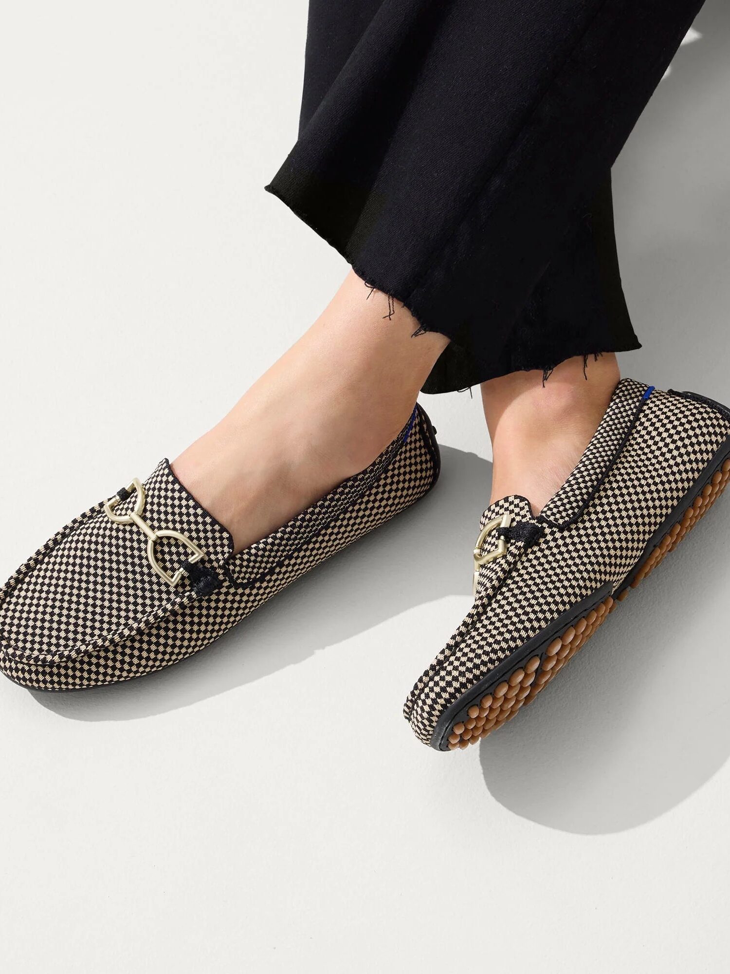 Person wearing black pants and checkered loafers with a decorative chain, standing on a light gray surface.