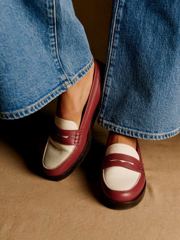 Person wearing flared blue jeans and maroon and white loafers standing on a beige surface.