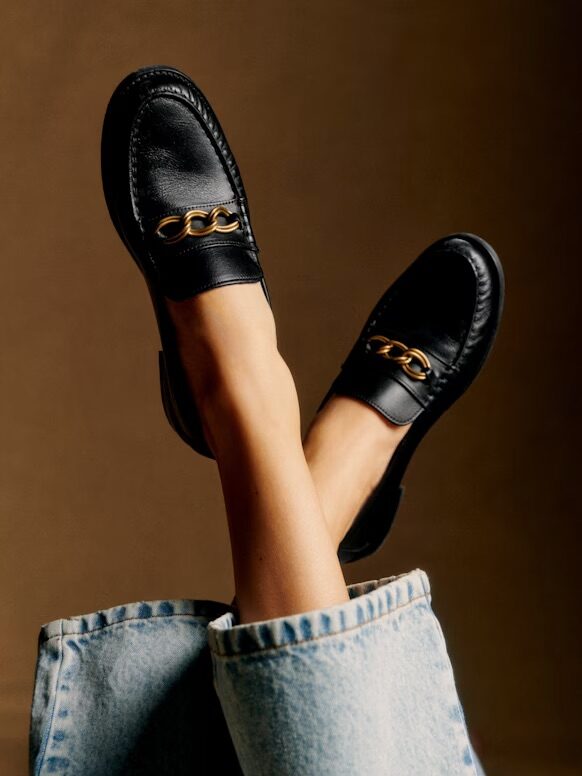 Person wearing black leather loafers with gold chain detail, feet crossed, and blue denim jeans visible against a brown background.