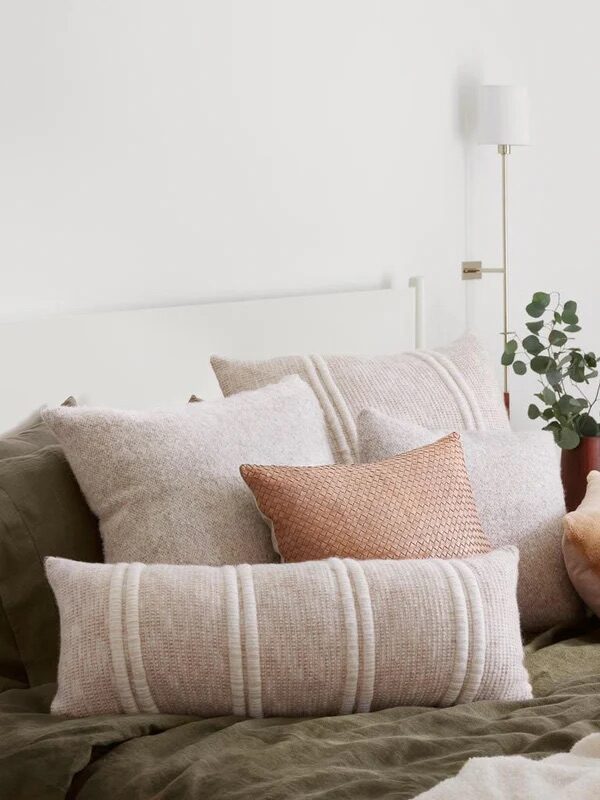 A neatly made bed with green bedding, five decorative pillows in neutral tones, and a small vase with greenery on the bedside table against a white wall.