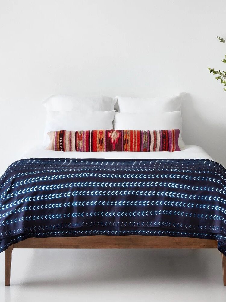 A neatly made bed with white pillows, a long decorative cushion with a red geometric pattern, and a dark blue blanket with a repeating white crescent design.