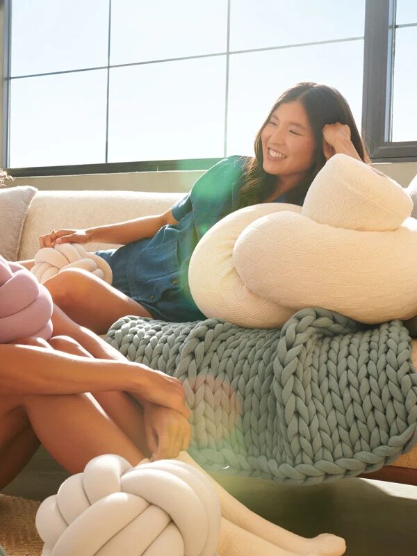 Two women sit on a sofa with bright sunlight, surrounded by chunky knit blankets and large knotted cushions, smiling and relaxing together.
