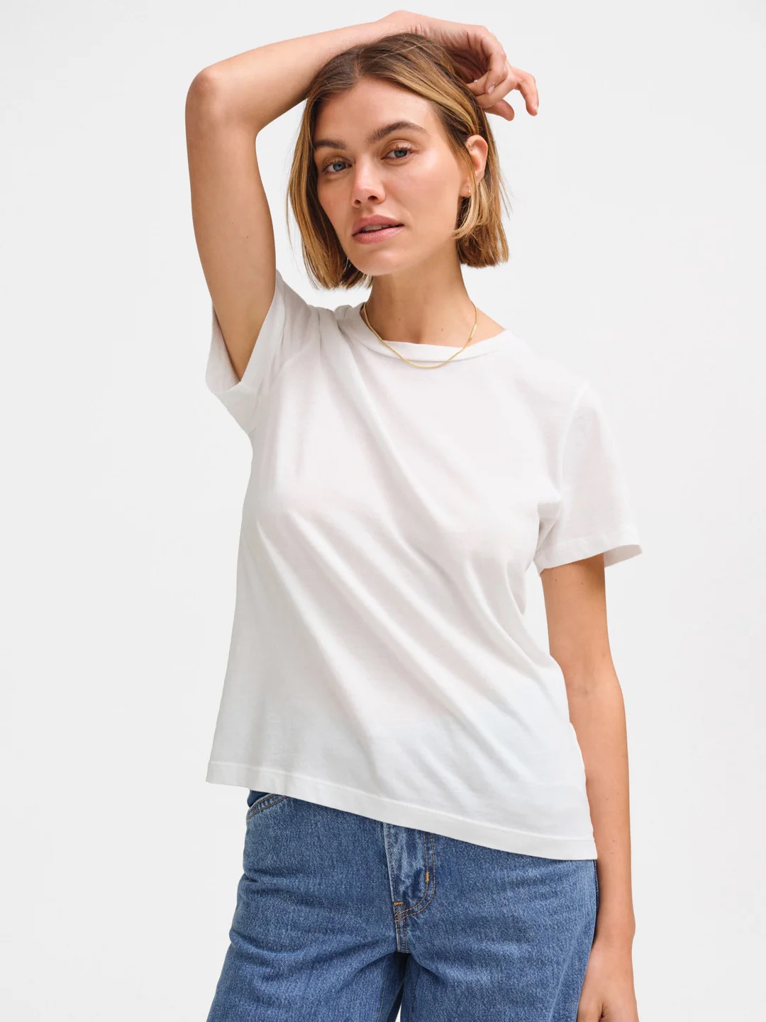 Person with short hair wearing a white T-shirt and blue jeans stands with one hand resting on their head, against a plain background.