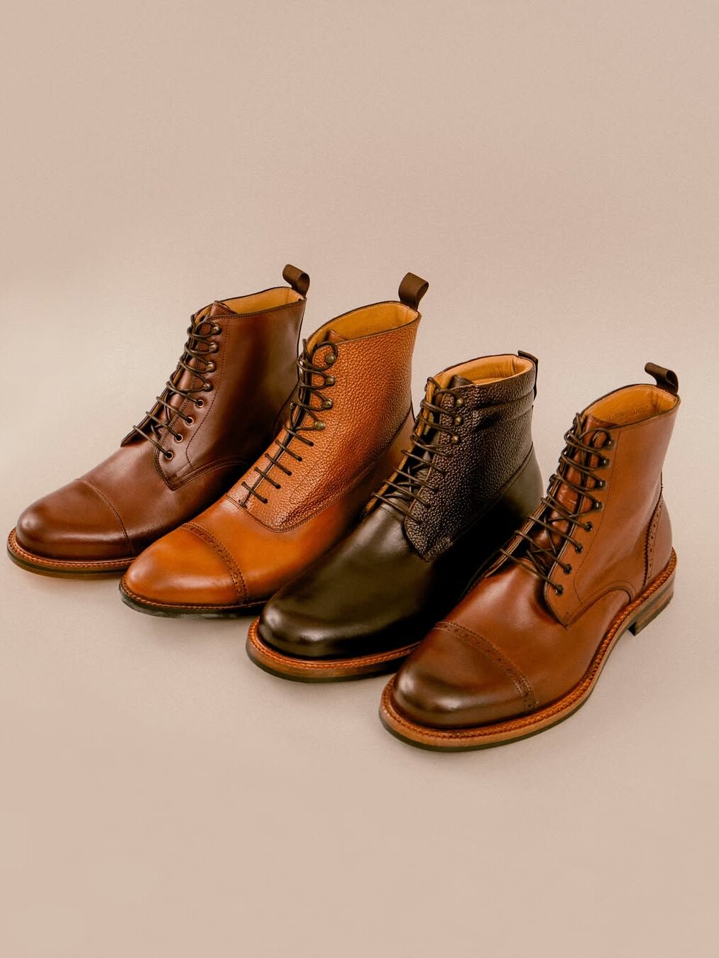 Four pairs of men's lace-up leather boots in different brown shades and textures are neatly arranged in a row on a beige background.
